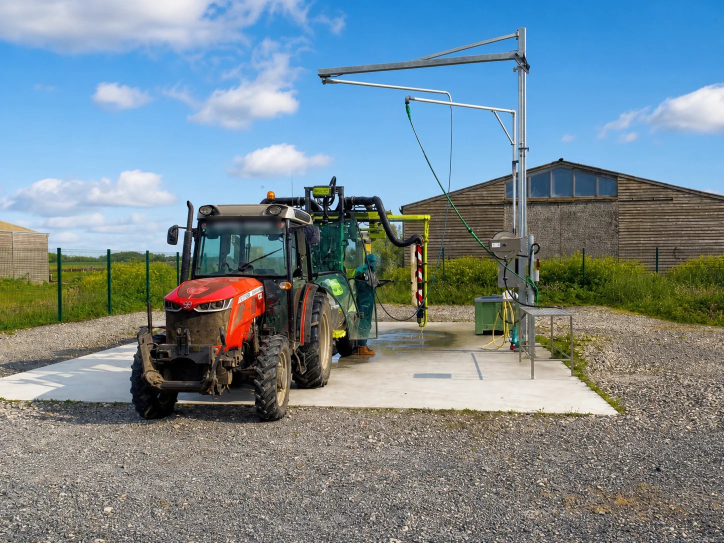 Tractor met veldspuit op een betonnen wasplaats met wasboom en hogedrukslang naast een landbouwschuur